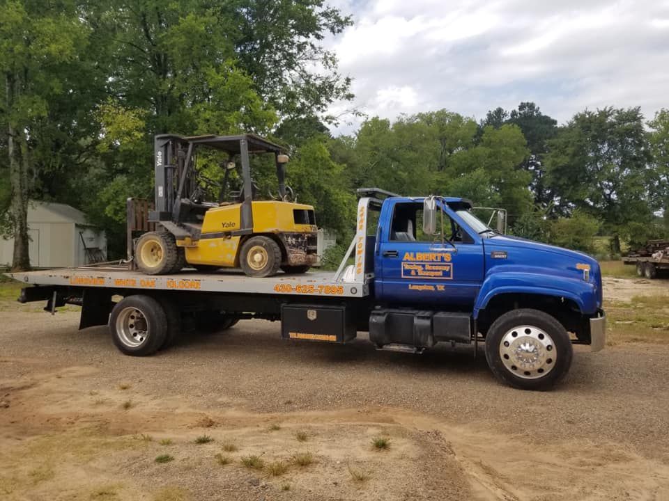 Blue tow truck carrying a yellow forklift in a rural setting.
