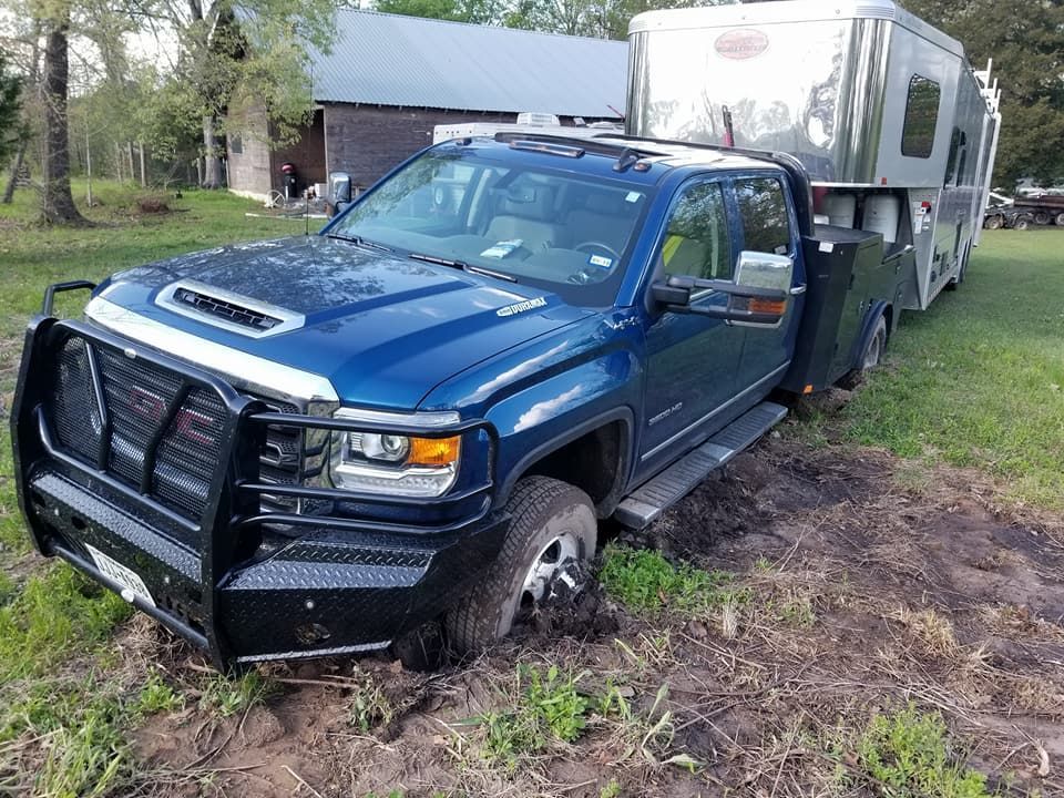 Blue truck stuck in mud, towing a horse trailer, in a grassy field near a barn.