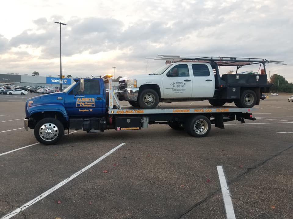Blue tow truck carrying a white work truck on a flatbed in a parking lot.