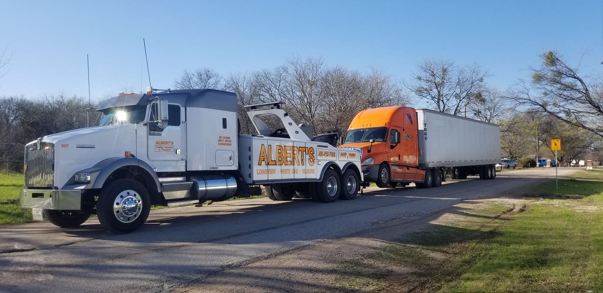 A tow truck pulling an orange and white semi-truck with a trailer on a road; a clear day.