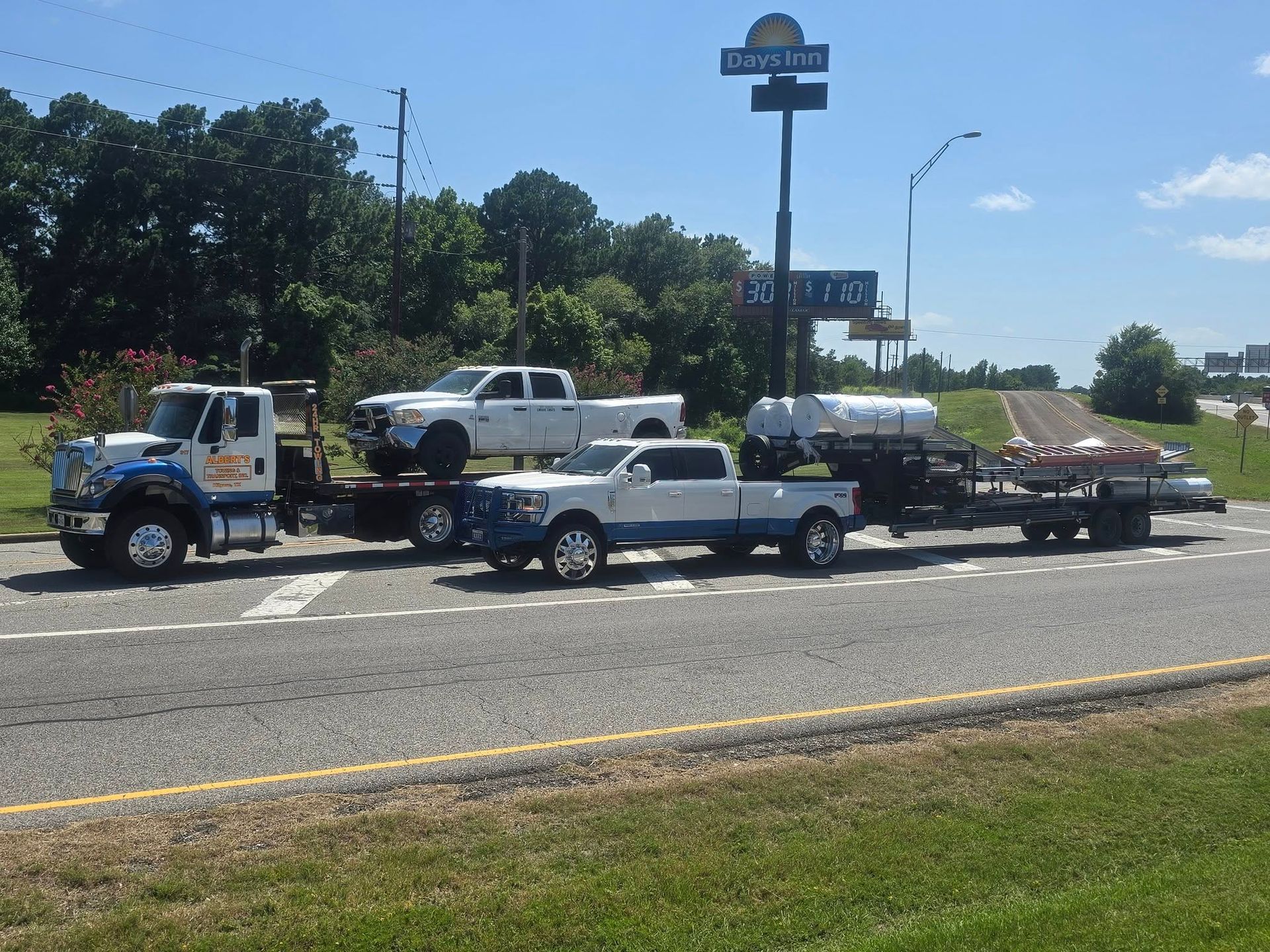 Tow truck hauling two trucks and a trailer with tanks, on a road in front of a Days Inn sign.