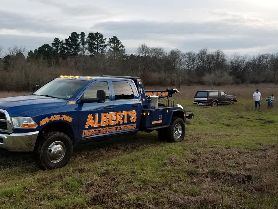 Blue Albert's tow truck in a field towing a vehicle. Two people watch nearby. Overcast sky.