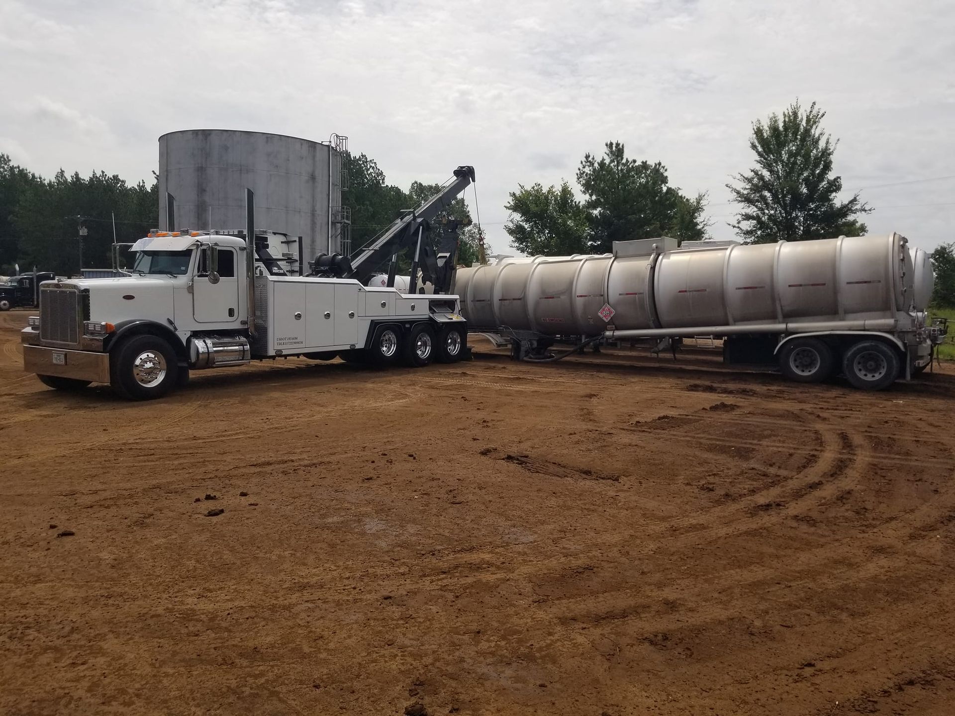 White tow truck pulling a silver tanker on a dirt field, with a silo in the background.