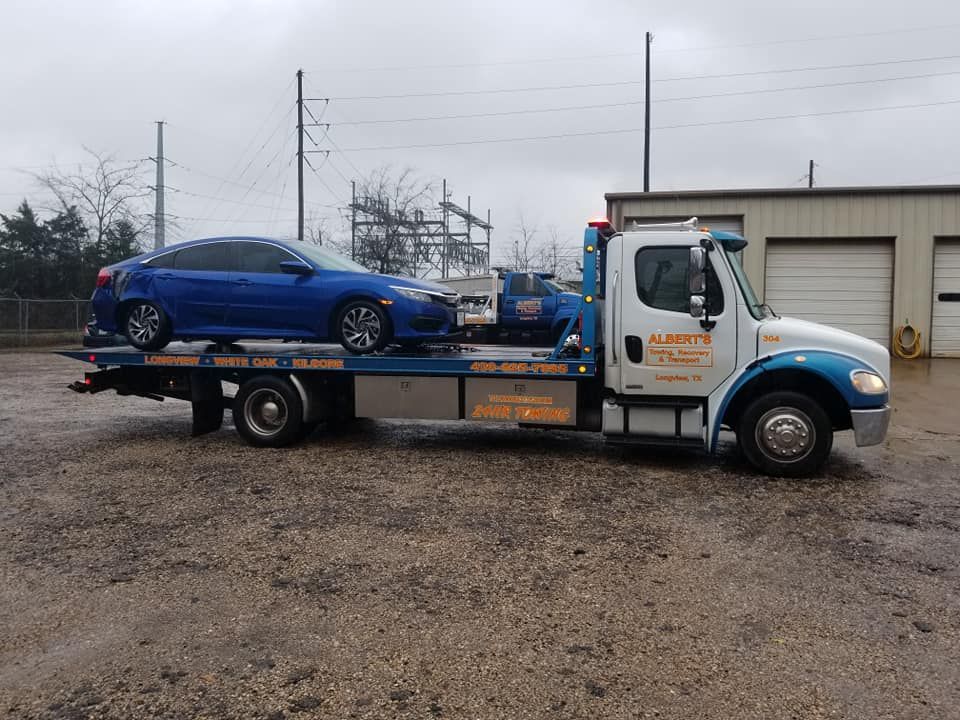 Blue car on a tow truck; parked outdoors, cloudy day. Truck is white and blue, with company logo.