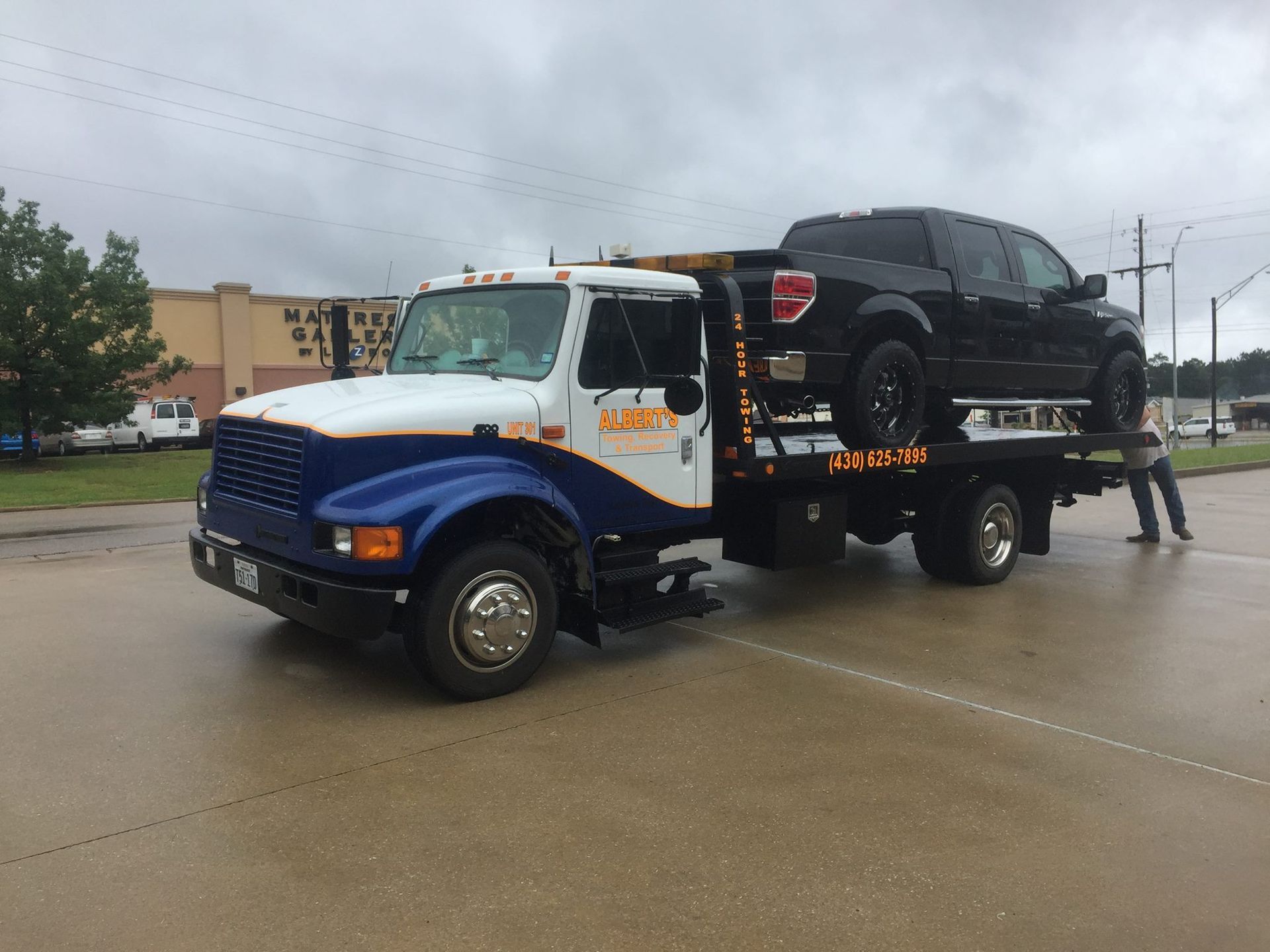 Tow truck hauling a black pickup truck on a wet road. Blue, white, and orange truck.