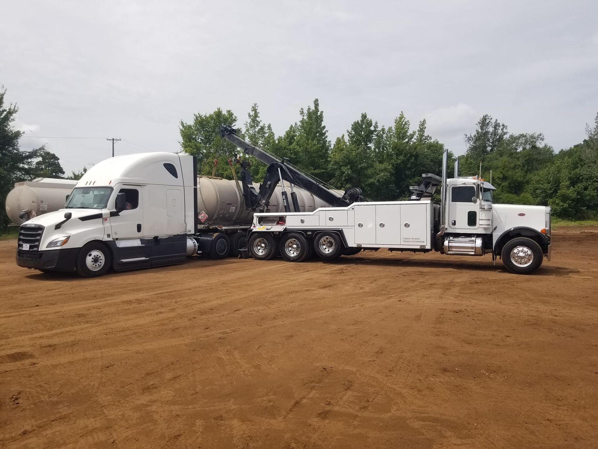White semi-truck being towed by a larger white tow truck on a dirt lot, trees in the background.
