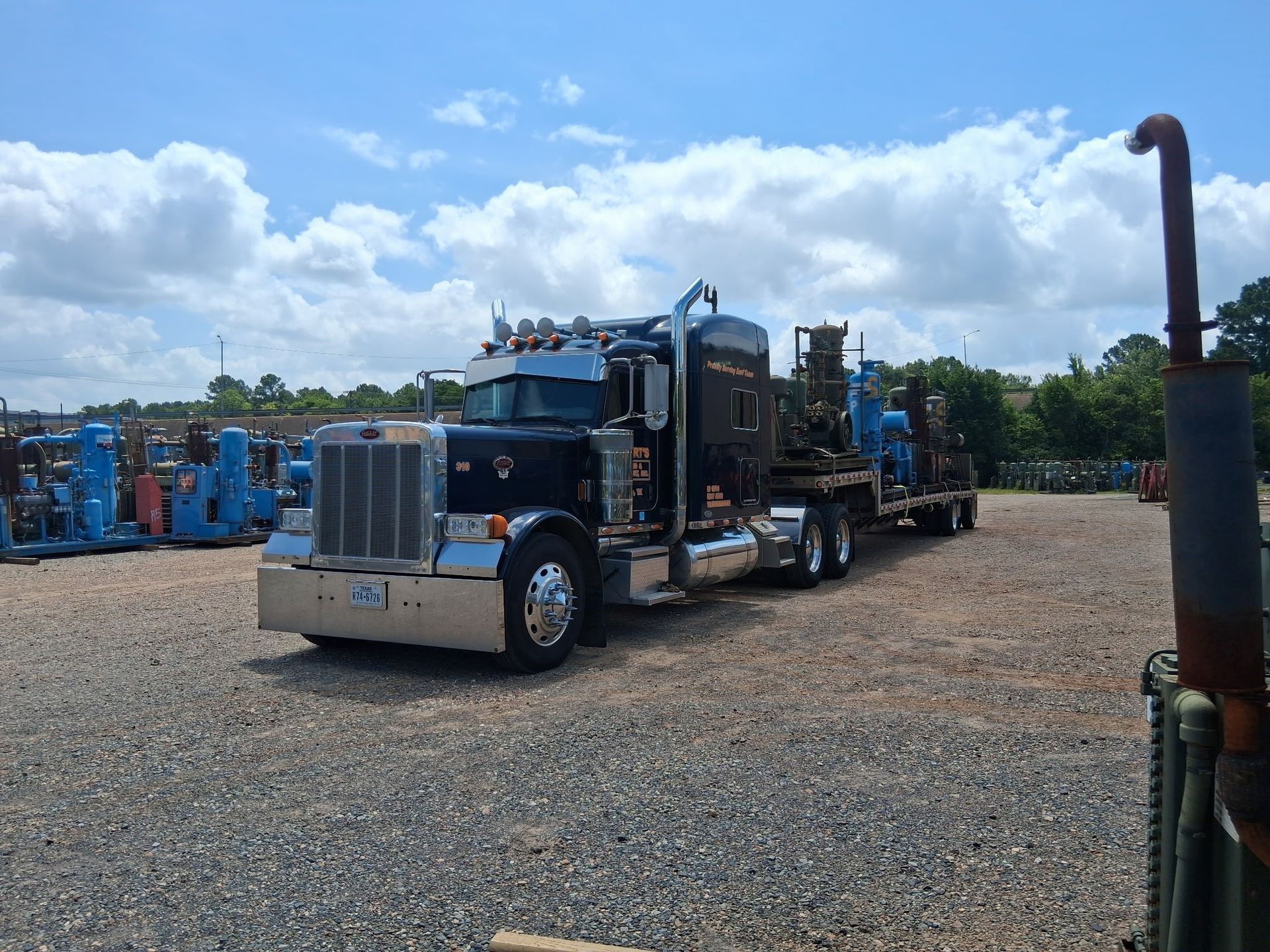 Black semi-truck with chrome accents hauling equipment on a trailer, parked on gravel under a blue sky.