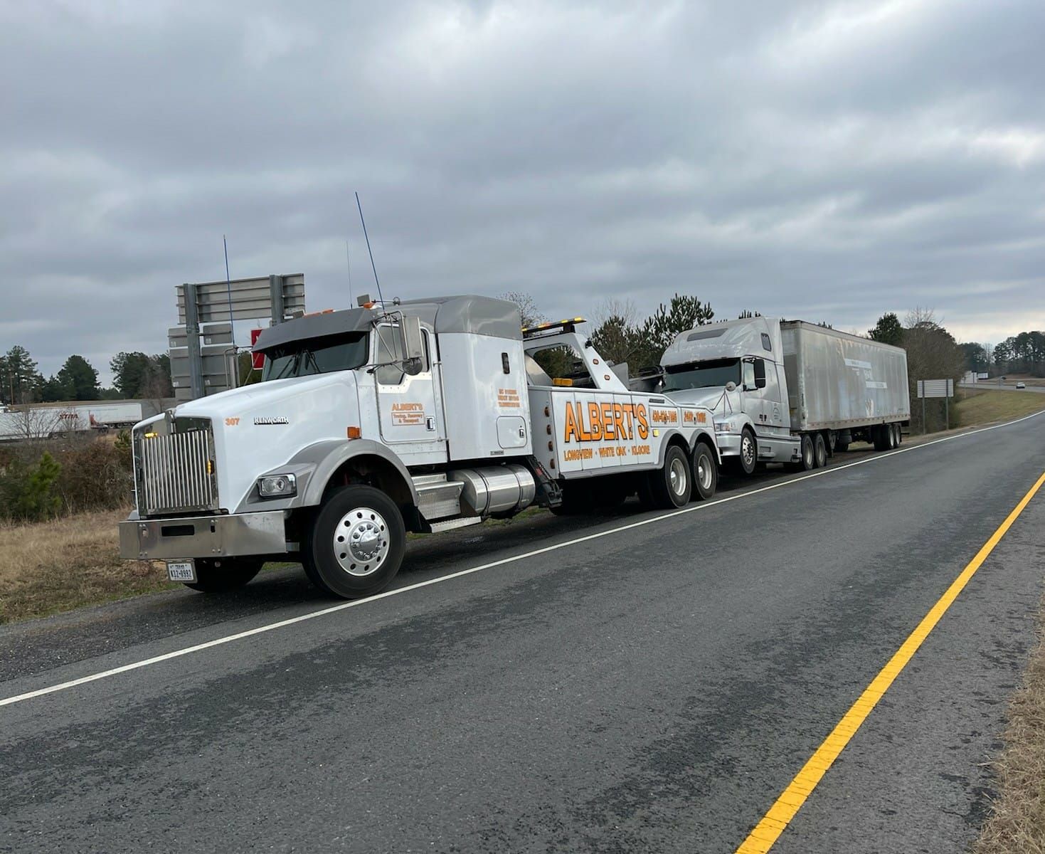 Tow truck towing another truck with a silver trailer on a roadside. Cloudy day.