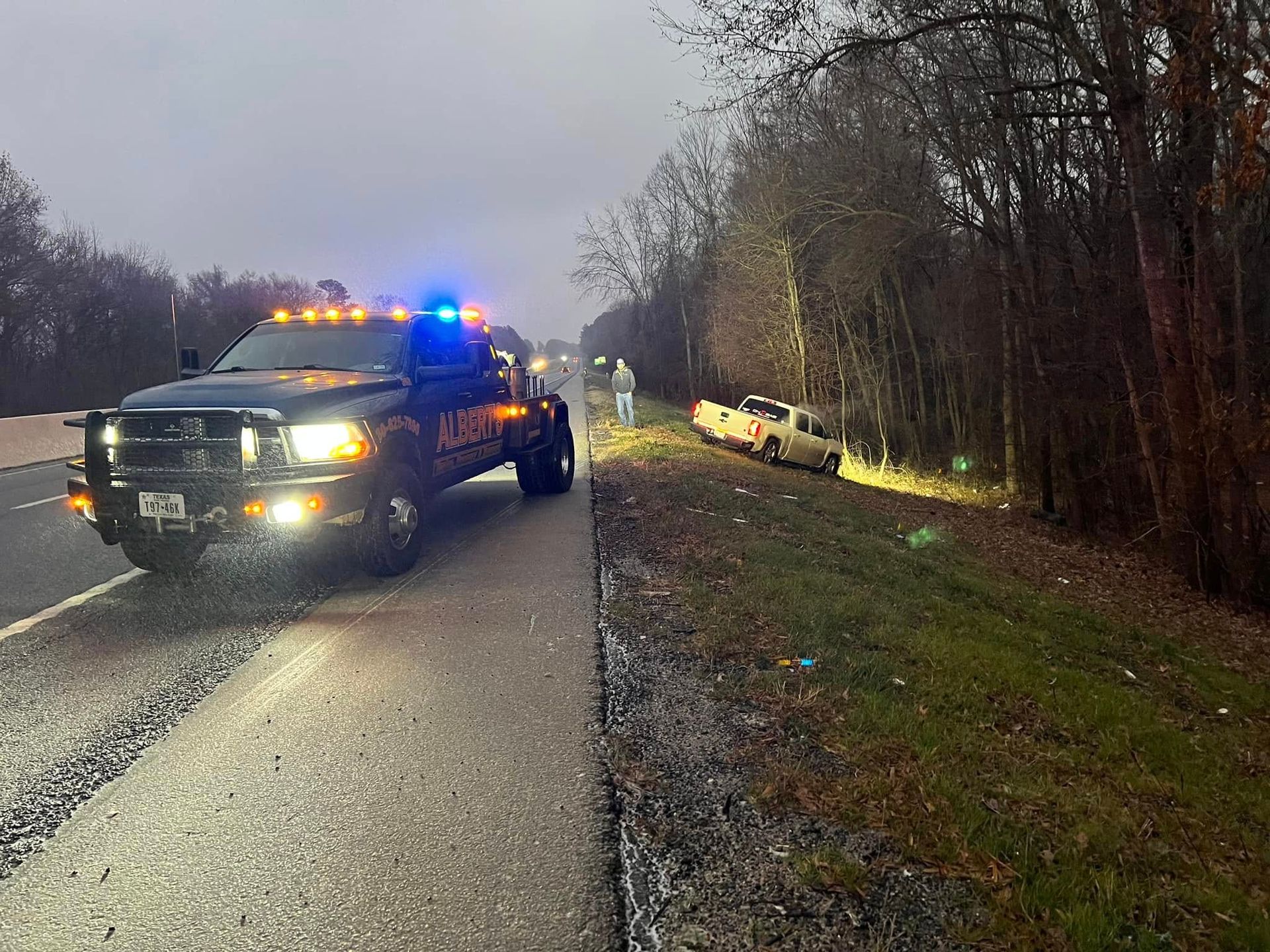 Tow truck on a highway assisting a white pickup truck in the ditch near a wooded area at dusk.