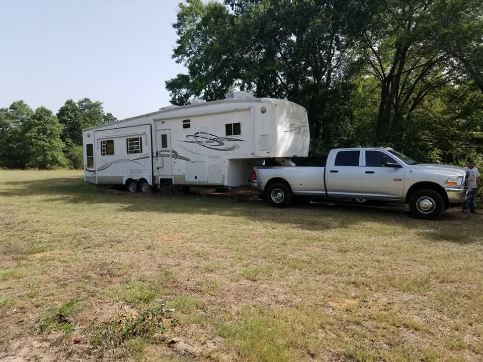 Silver pickup truck towing a white fifth-wheel RV in a grassy field on a sunny day.