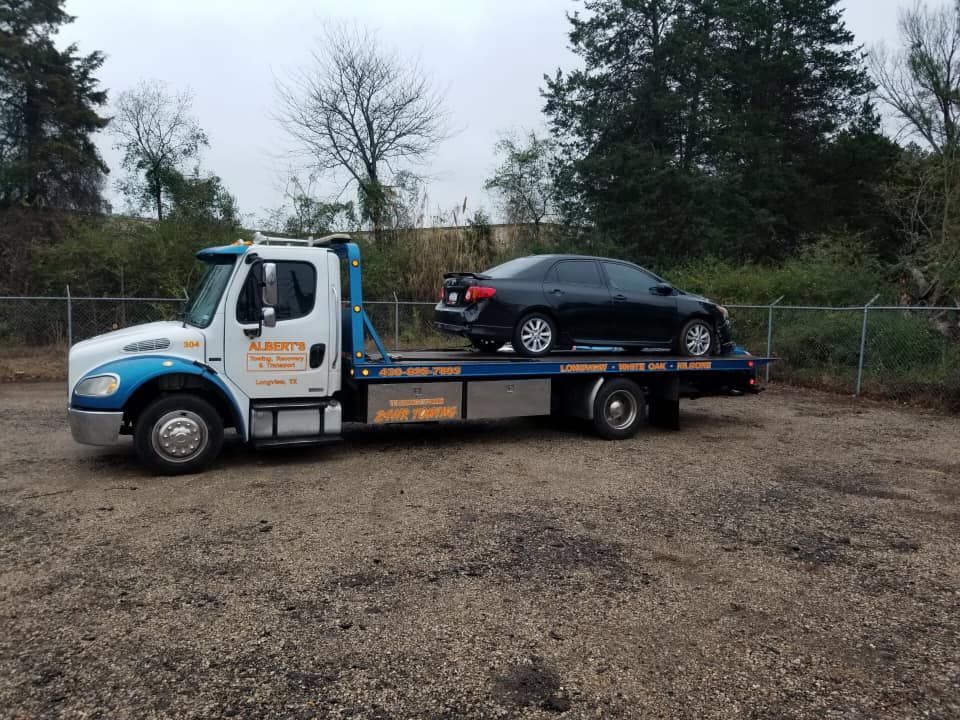 Tow truck hauling a black car on a flatbed; parked in a gravel lot, trees in the background.
