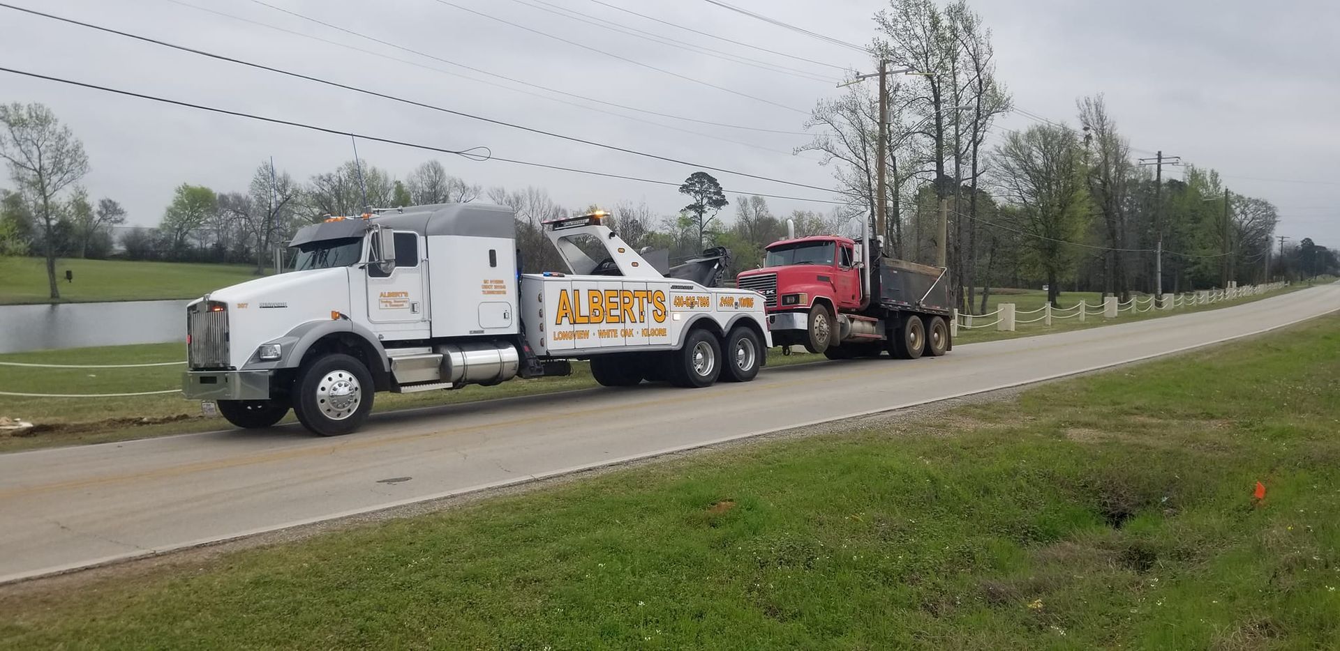 A white tow truck towing a red dump truck down a paved road, grass on either side, under power lines.