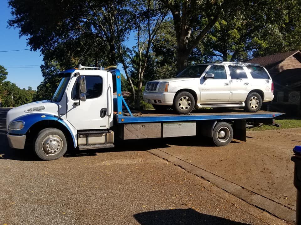 Tow truck with white Cadillac SUV loaded on the flatbed in a residential area.