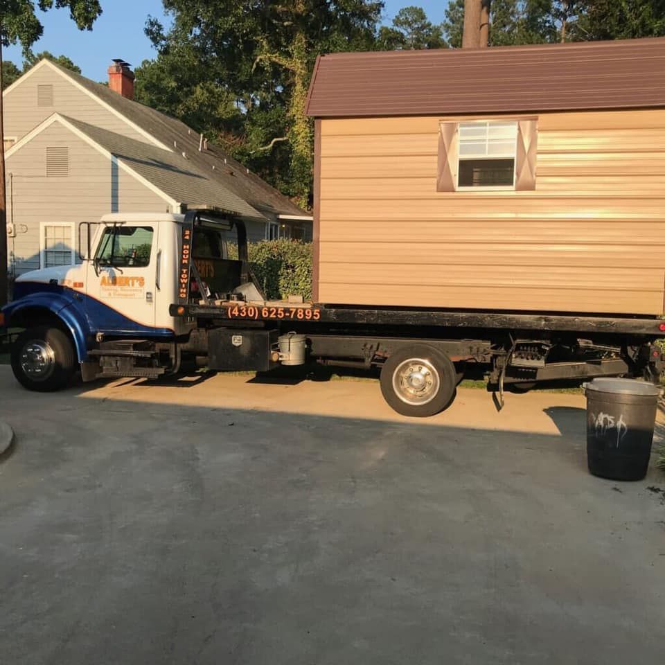 A tow truck carrying a brown shed on a flatbed, parked in a driveway.