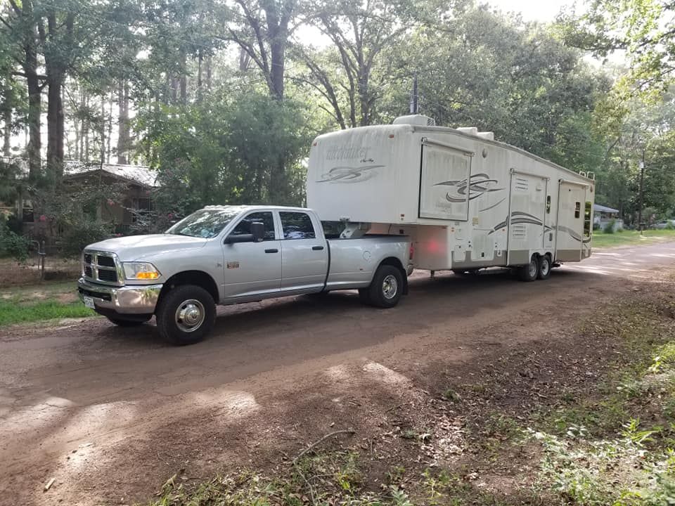 Silver pickup truck towing a large, white RV on a dirt road in a wooded area.
