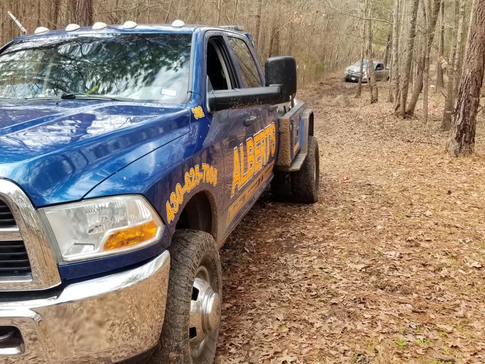 Blue tow truck on a dirt road in a wooded area, potentially rescuing another vehicle.