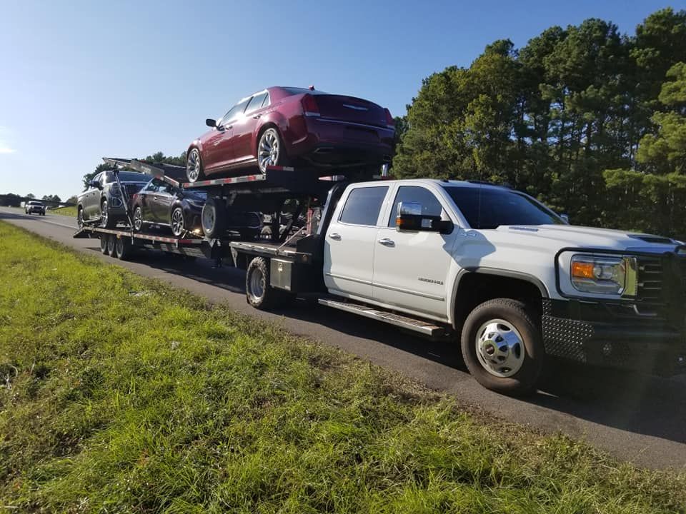 White truck hauling a trailer with several cars on a roadside with green grass and trees.