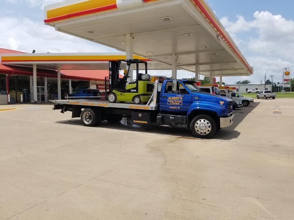 Blue tow truck with a green forklift on its flatbed at a Shell gas station.