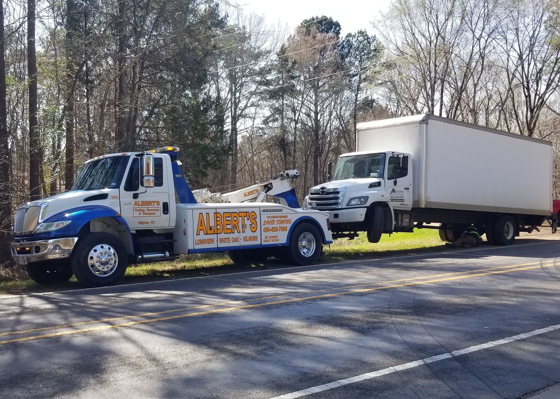 Tow truck towing a box truck on a road, trees in the background.