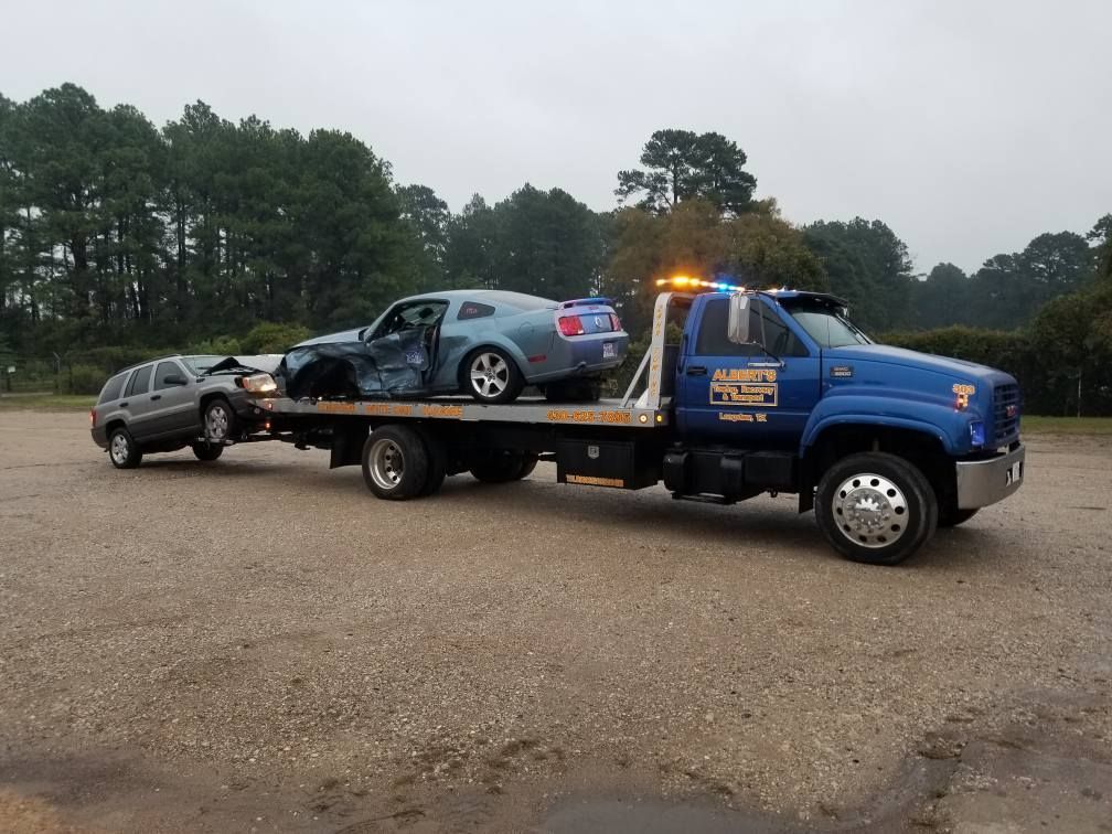 A blue tow truck carrying a damaged blue car and a silver SUV on a gravel lot.