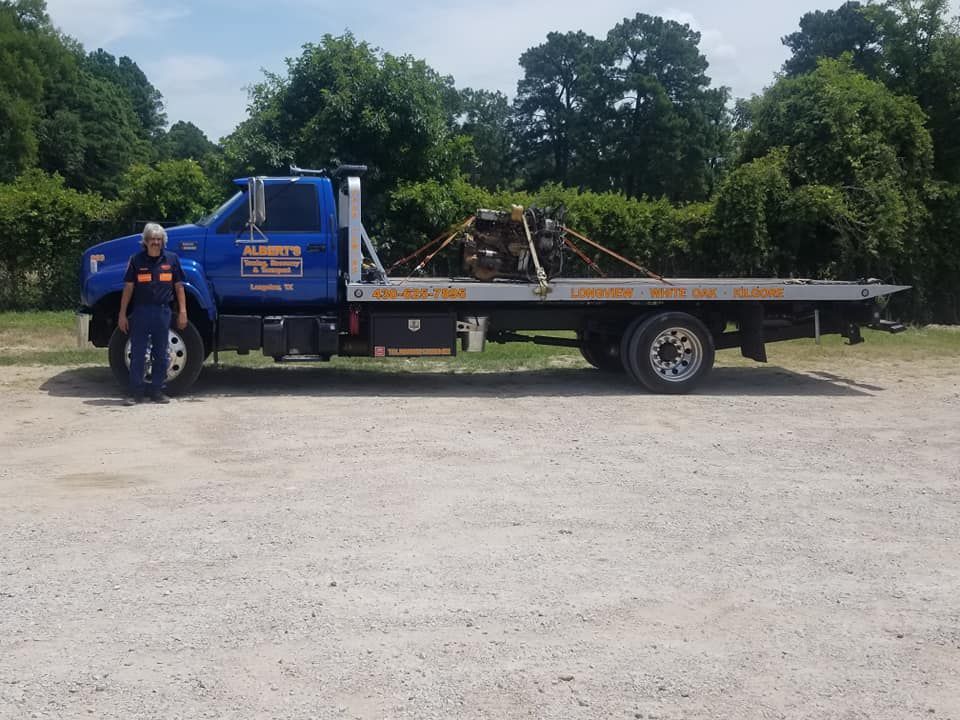 Woman in uniform stands beside a blue tow truck on gravel. Trees in the background.