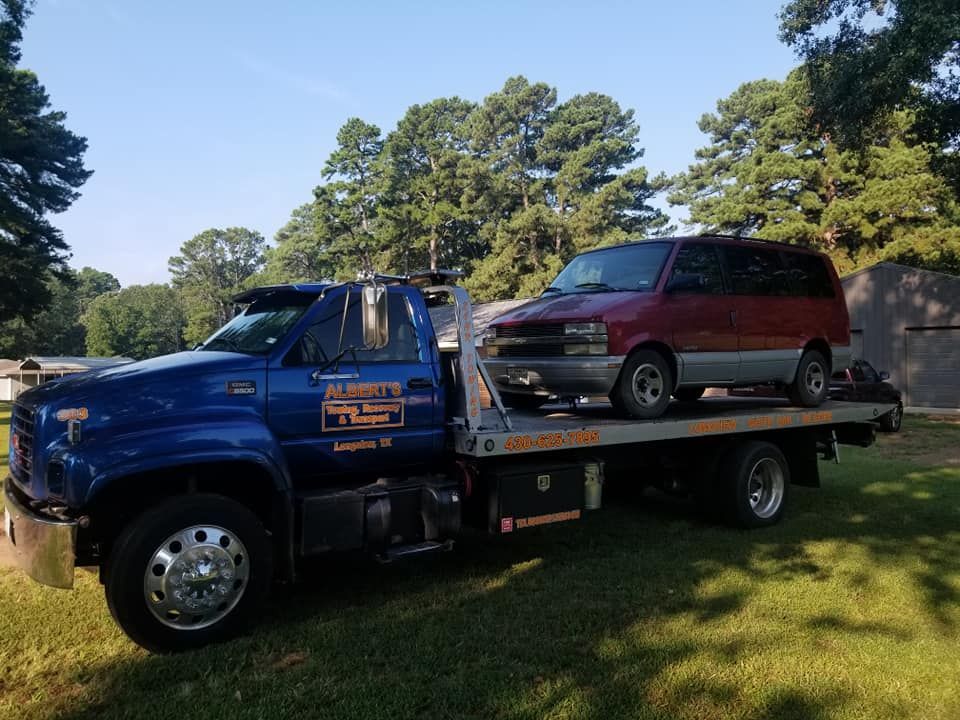 A blue tow truck carries a red minivan on its flatbed in a grassy outdoor setting.