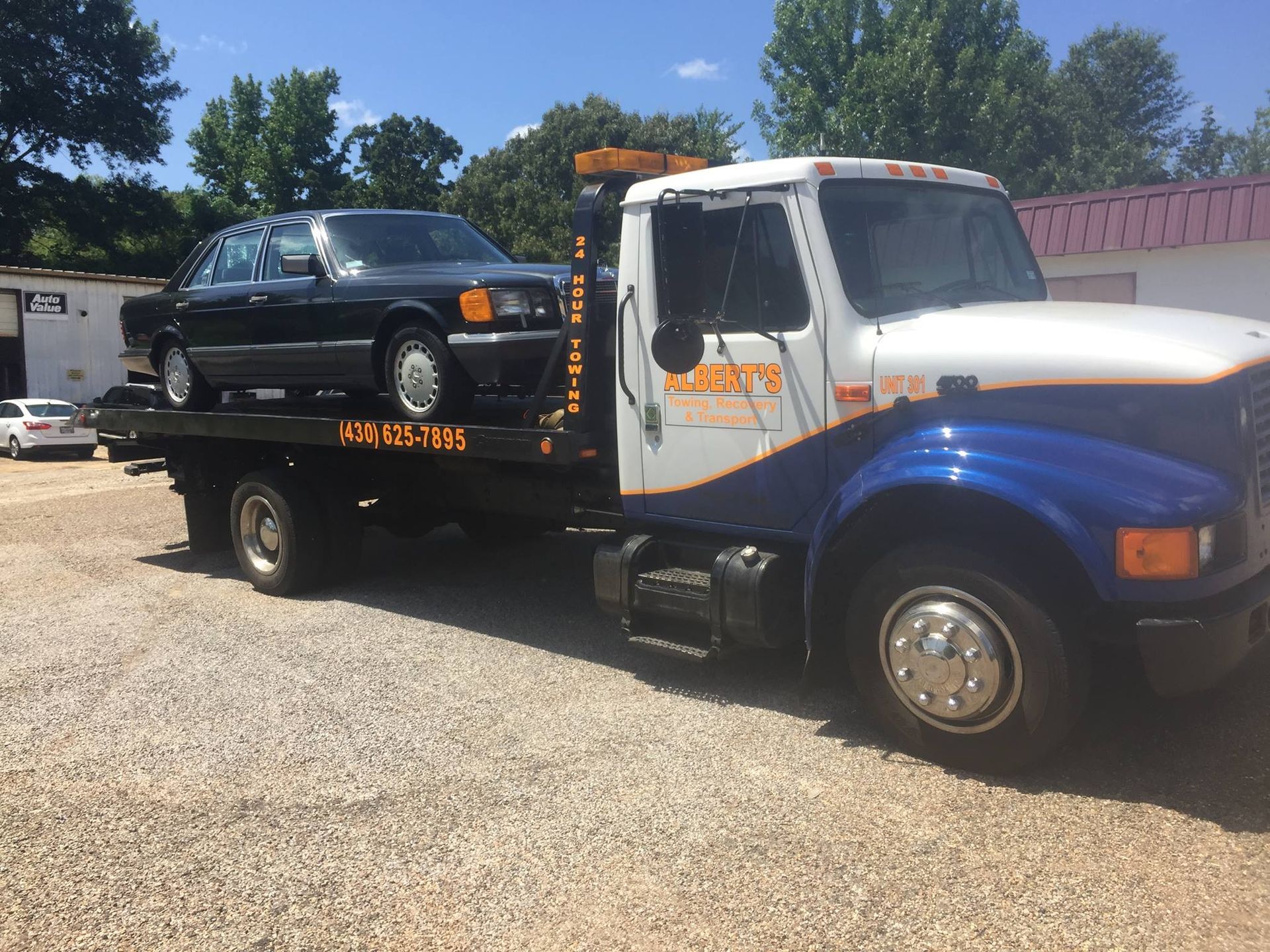 A black car on a tow truck, white and blue cab, parked on gravel in front of a building.