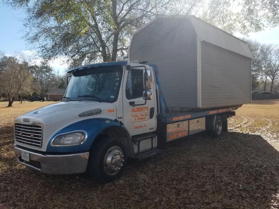 A blue and white tow truck transporting a grey shed on its flatbed in a grassy area.