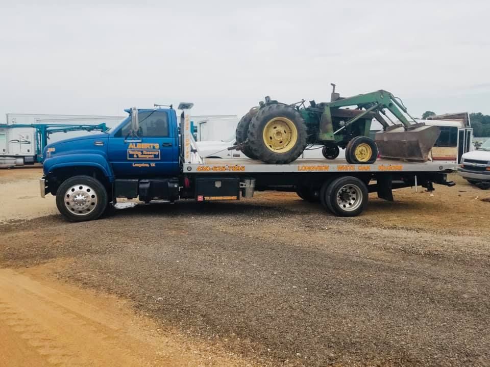 Blue tow truck carrying a John Deere tractor on a gravel lot.
