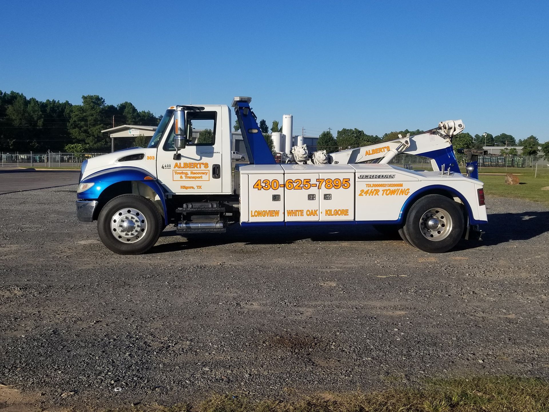 White and blue tow truck parked on gravel, under a clear blue sky. Phone number on the side.