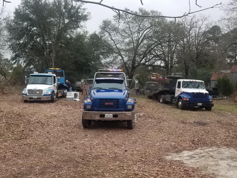 Three tow trucks parked on a leaf-covered lot. Blue and white trucks with trees in the background. Overcast sky.