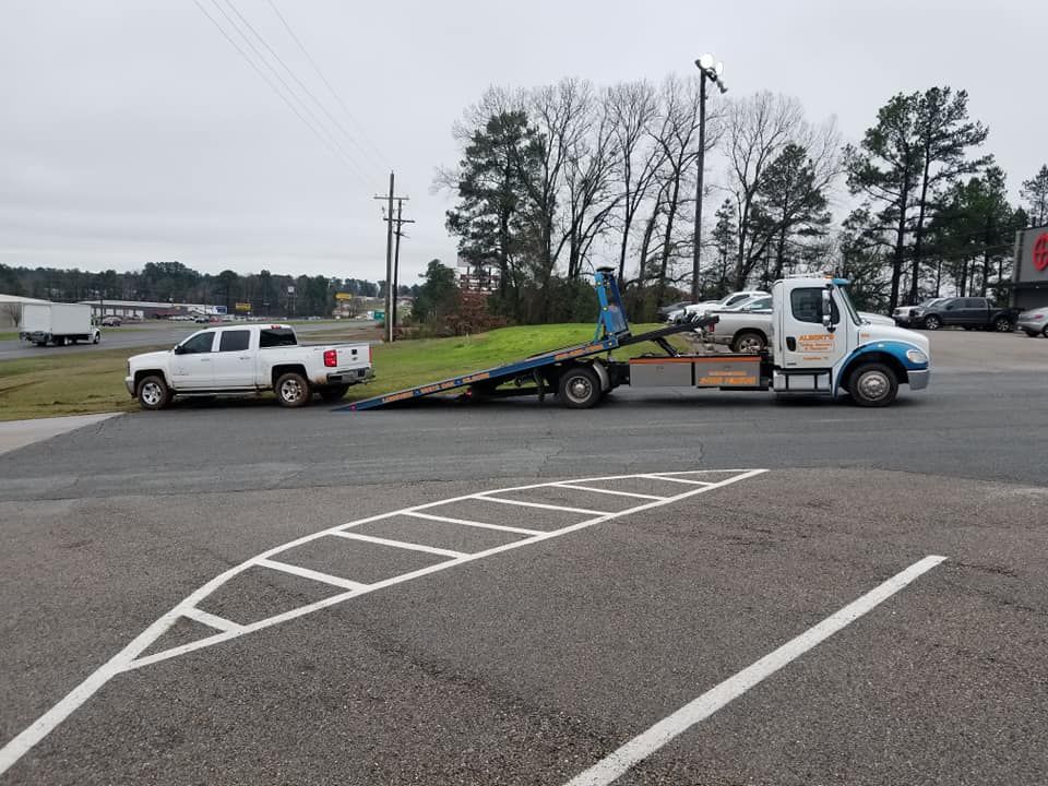 A white tow truck towing a white pickup truck on a flatbed trailer in a parking lot.