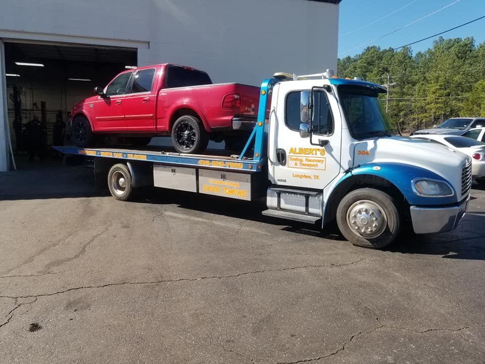 Red pickup truck being towed by a flatbed truck outside a repair shop.