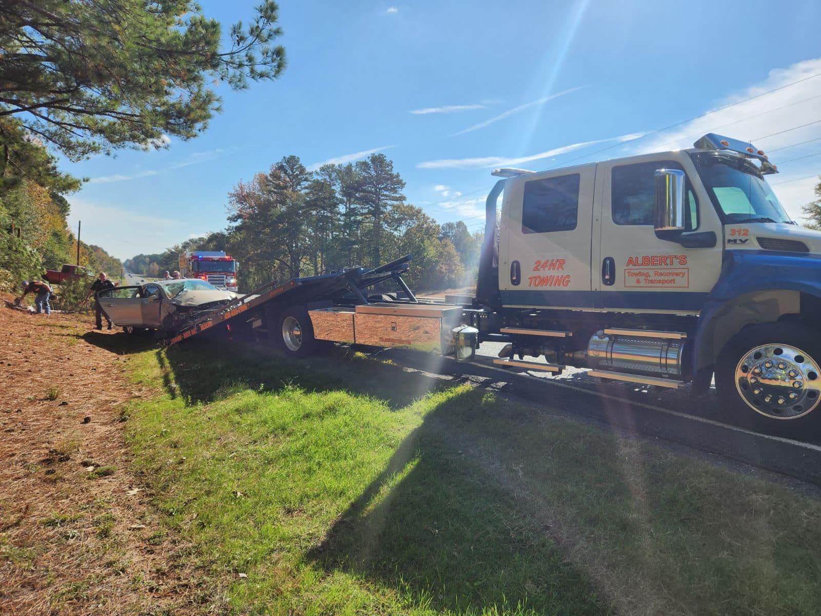 Tow truck loading a damaged silver car on a roadside, sunny day.