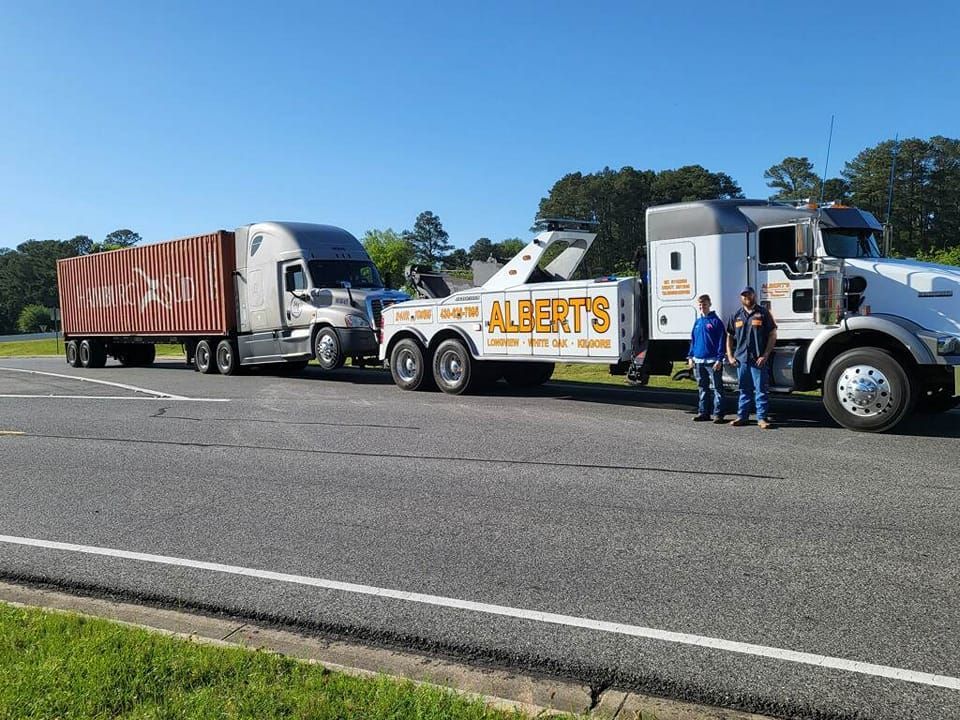 A large tow truck towing a semi-truck with a cargo container. Two people stand nearby on a sunny road.