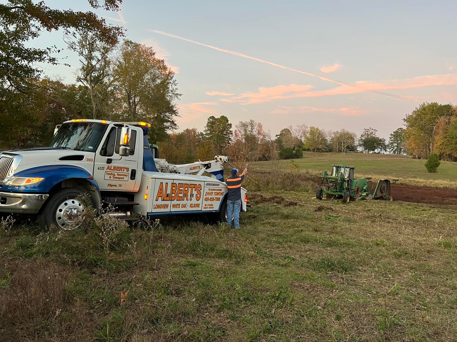 Tow truck with a person assisting a tractor in a field; sunset.