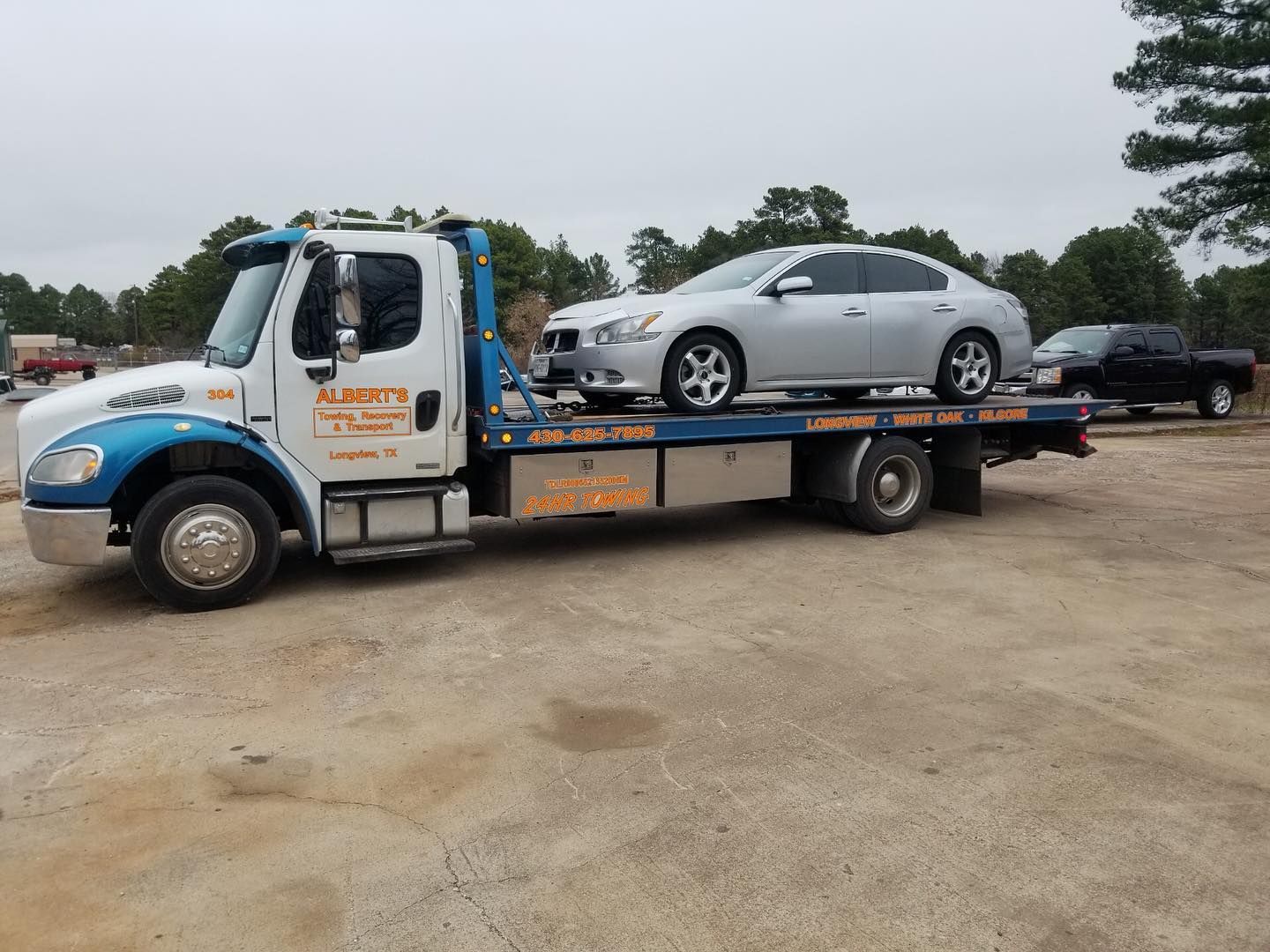 Tow truck carrying a silver sedan on a flatbed in a parking lot under an overcast sky.