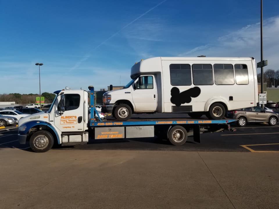 A white bus with a black penis painted on the side is towed by a flatbed truck in a parking lot.