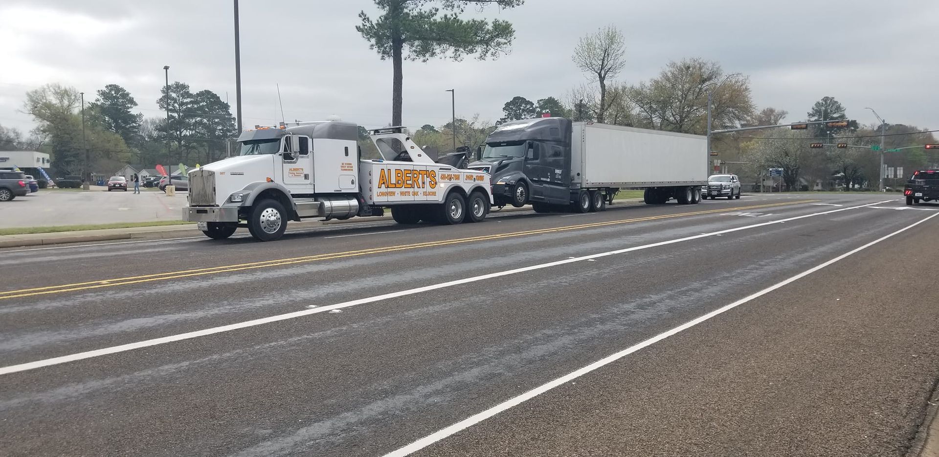 Tow truck towing a semi-truck on a road, overcast day. The tow truck has white and yellow colors.