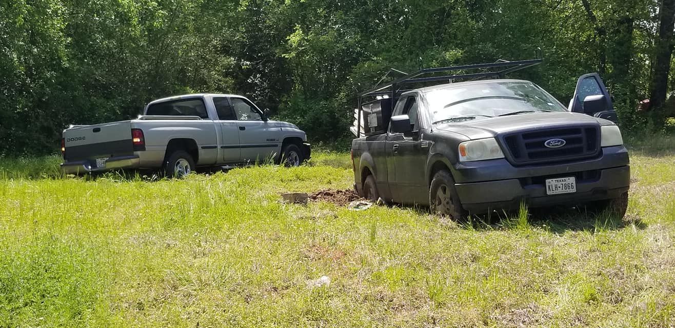 Two trucks stuck in mud in a grassy field near a tree line. The black truck has an open door.