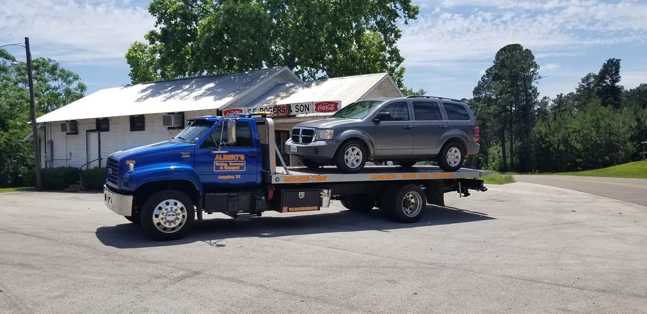 A blue tow truck carrying a gray SUV, in front of a building on a sunny day.
