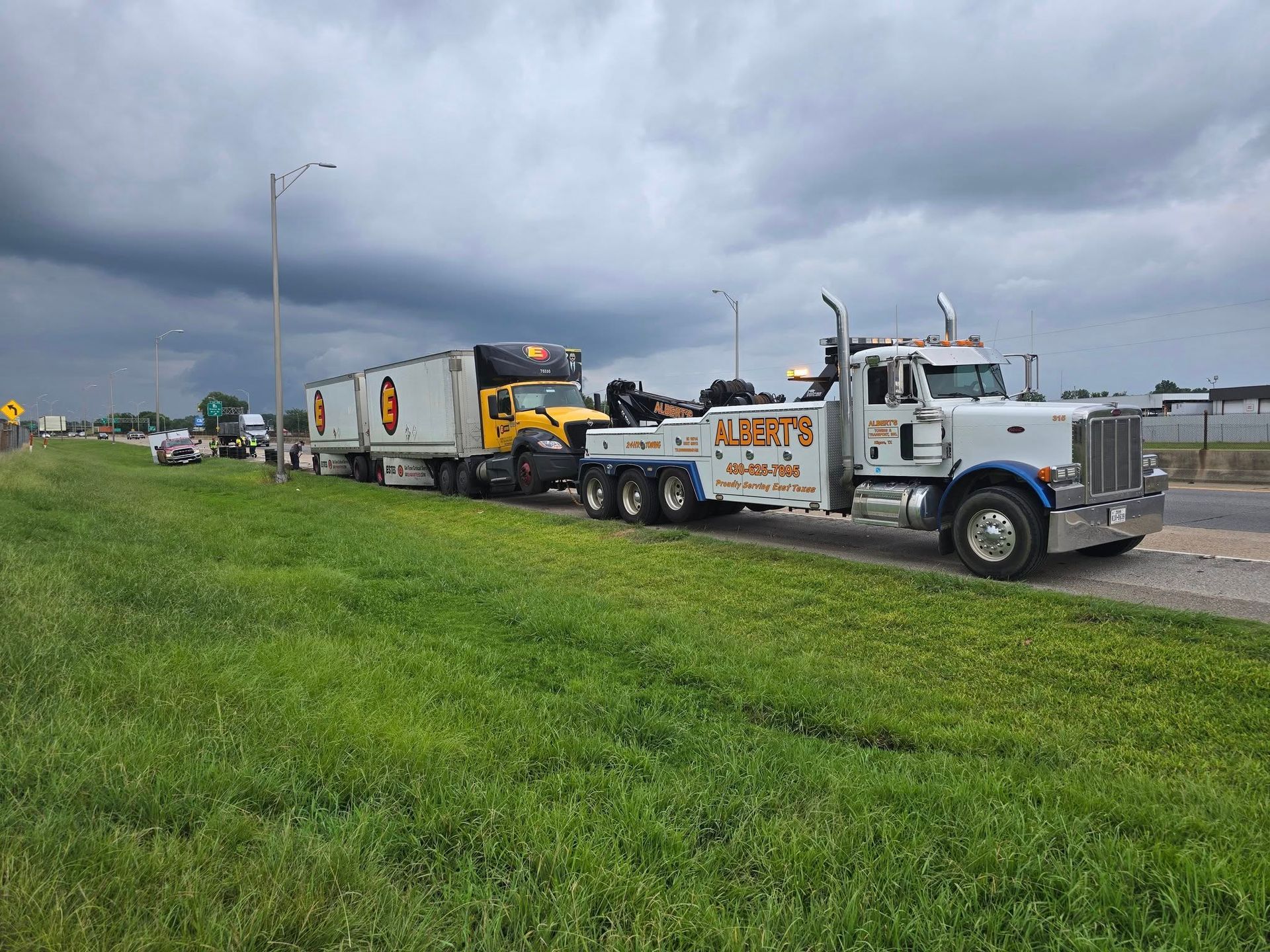 A semi-truck towing a UPS truck along a roadside with a cloudy sky overhead.