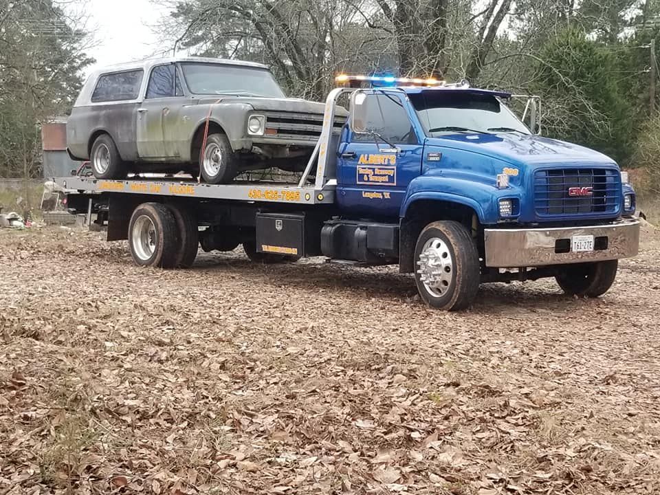 A blue tow truck carries a gray vintage SUV on a flatbed in a field.