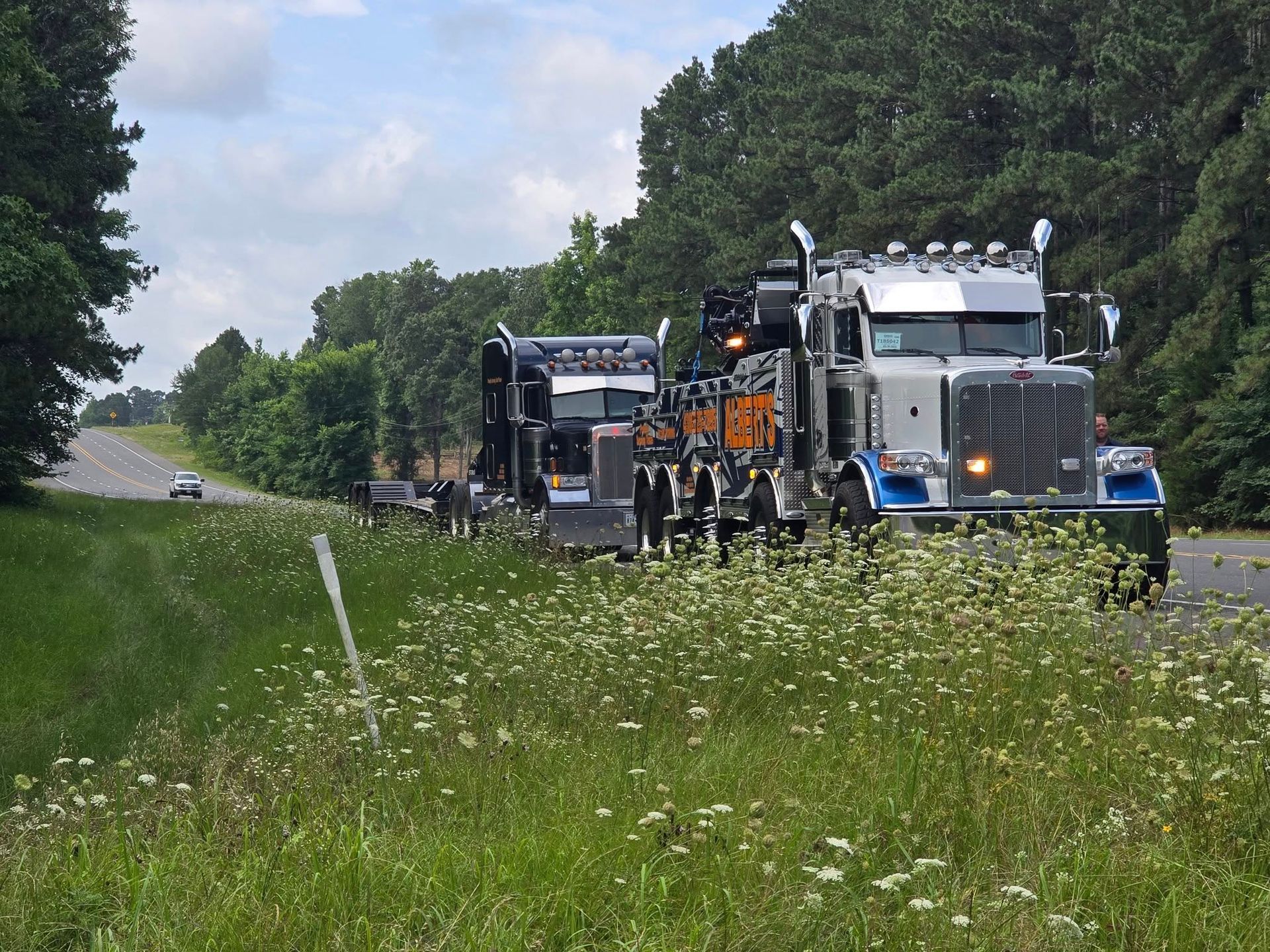 Semi-truck towing another semi-truck on a highway, roadside with tall grass and trees.