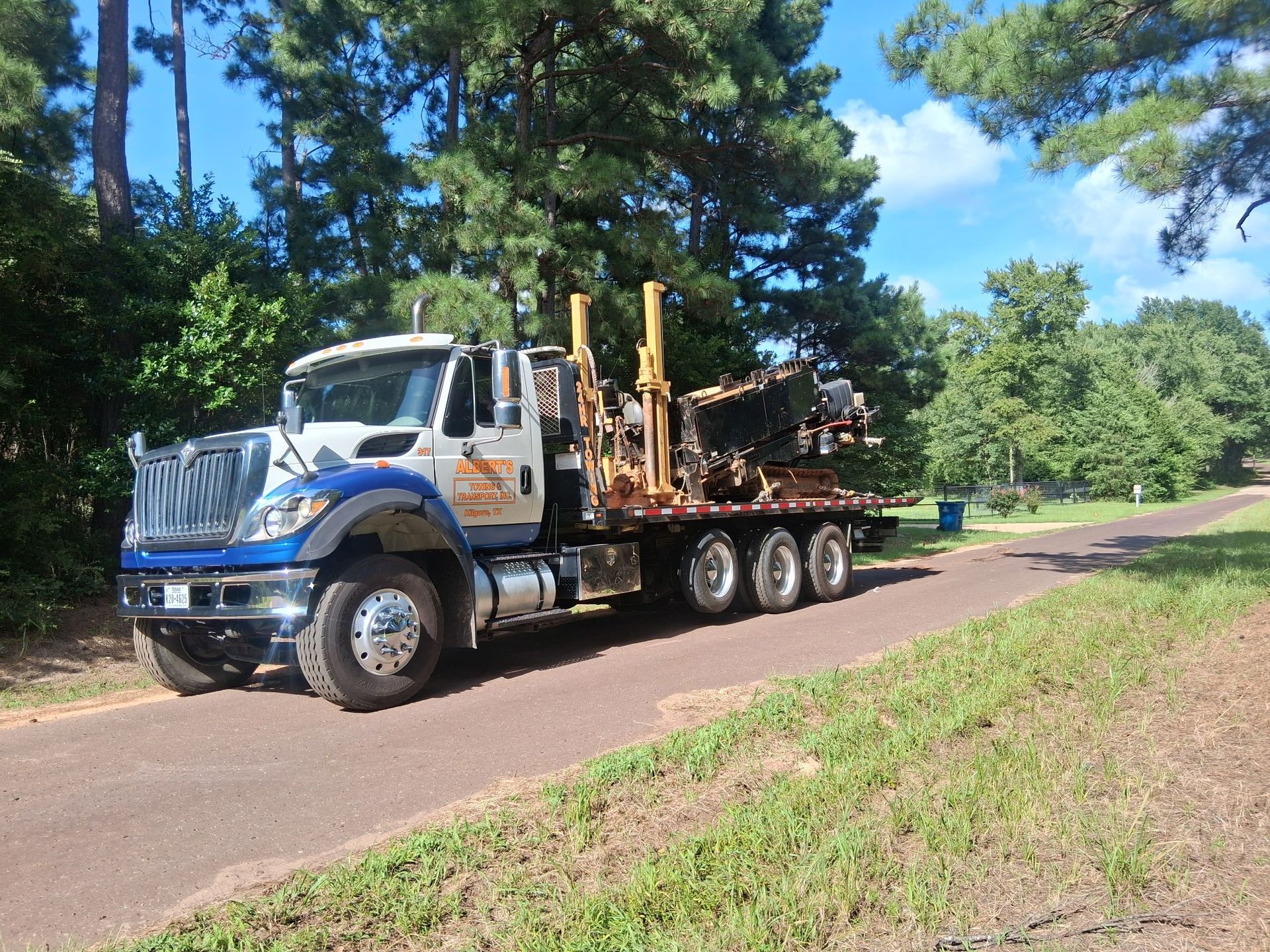 Logging truck on a road, blue and white cab, carrying logs. Trees and grass in background.