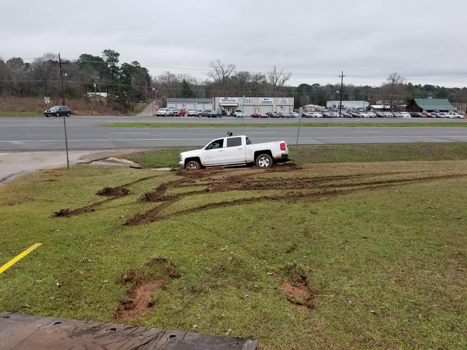 White pickup truck stuck in muddy grass next to a road, with tire tracks visible.