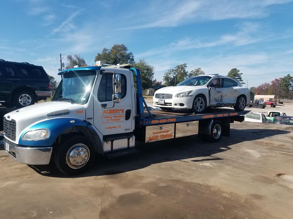 Tow truck with damaged white car, outdoors. Truck is white/blue with company logo; sunny day.