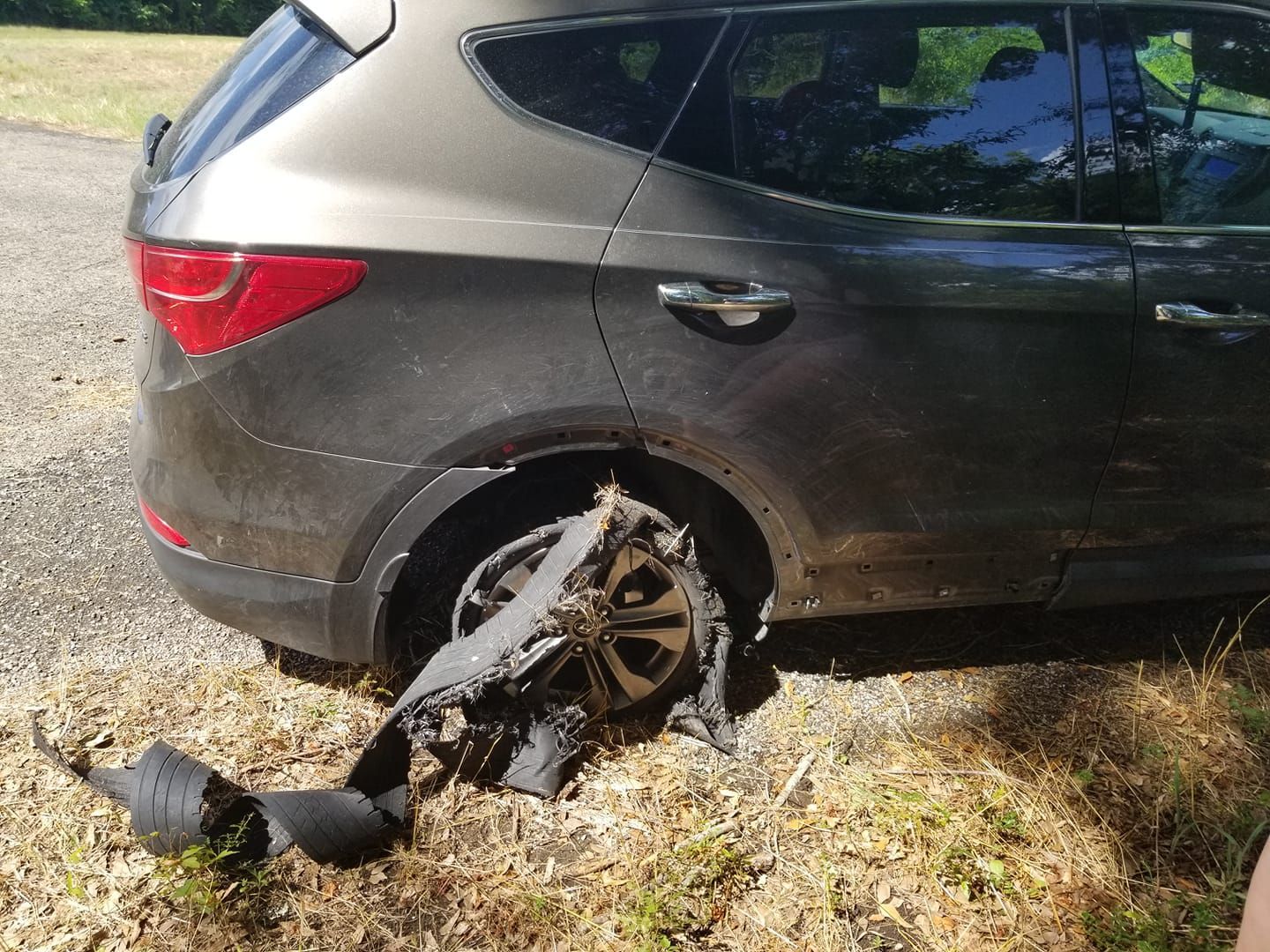 Gray SUV with a destroyed rear tire on a gravel road.