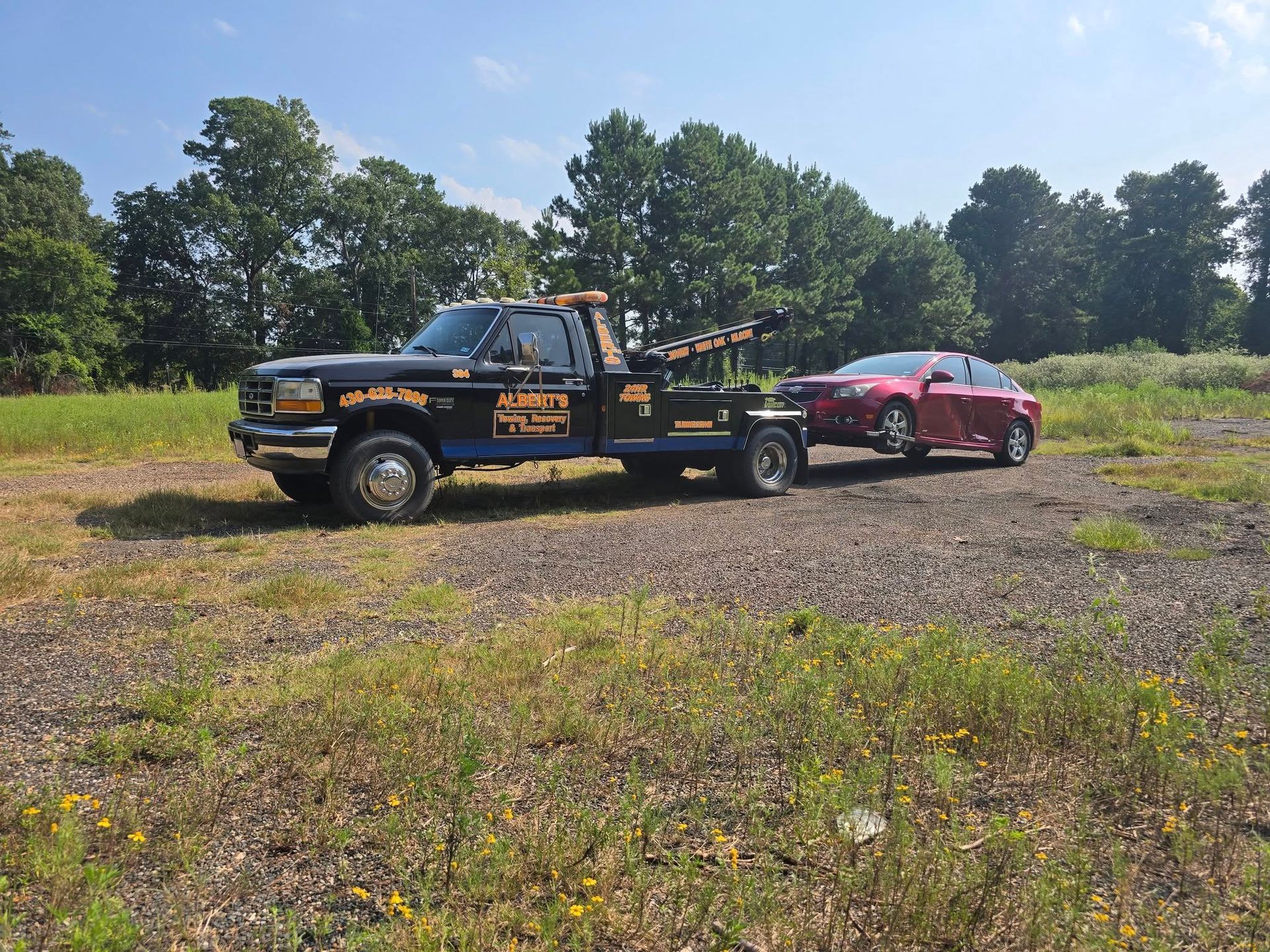 Tow truck towing a red car on a sunny day in a grassy field with trees.