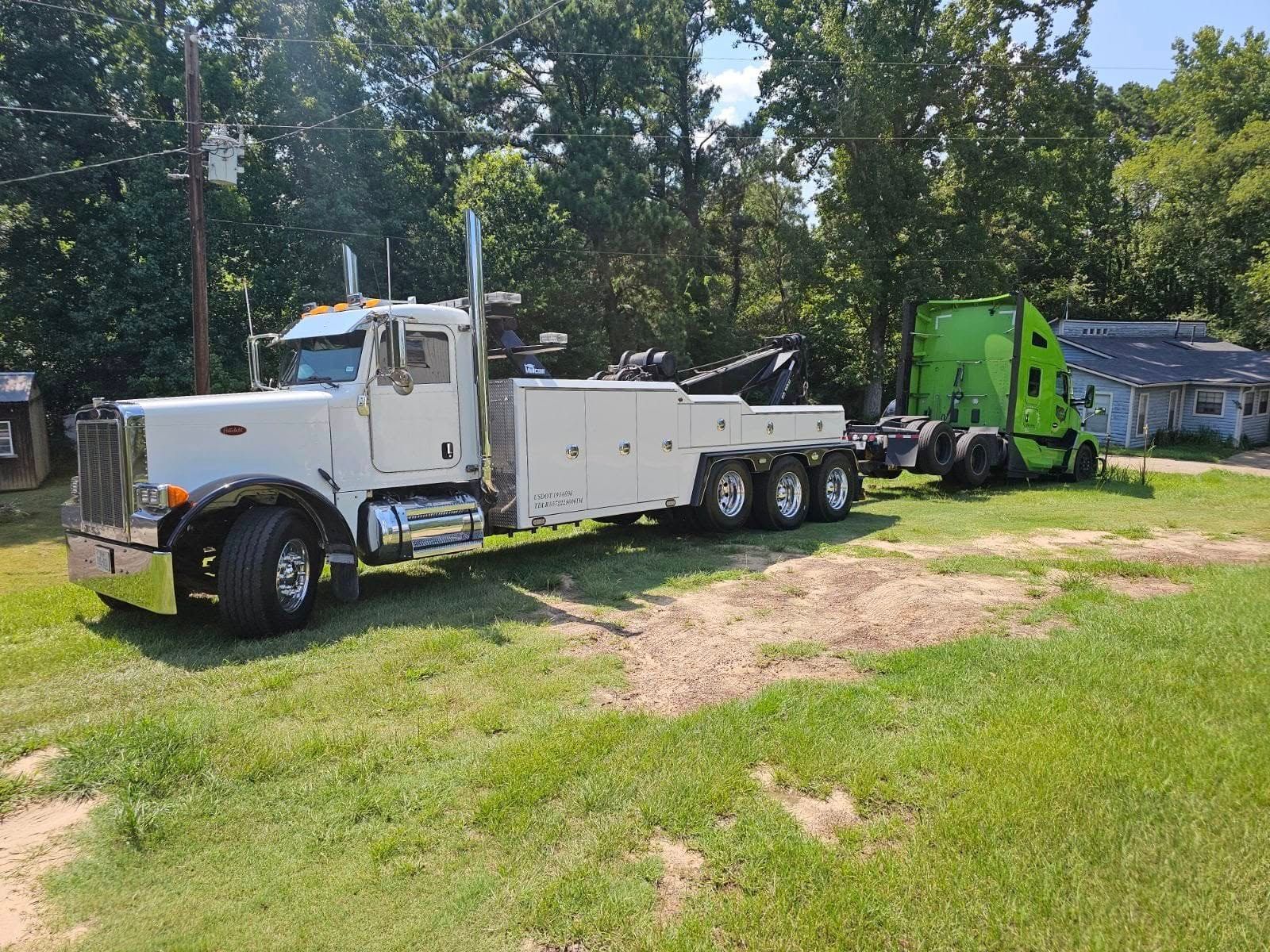 White tow truck towing a bright green semi-truck on grassy terrain, trees in background.